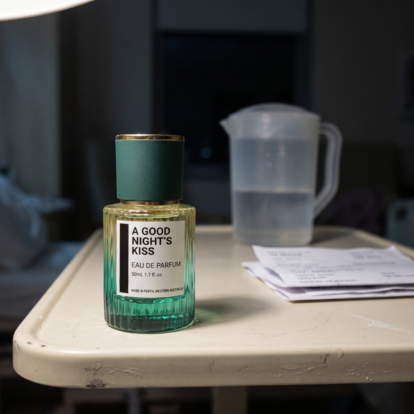 A fluted emerald glass perfume bottle labelled A Good Night's Kiss, standing on a hospital bedside table beside a water jug and a stack of paperwork.